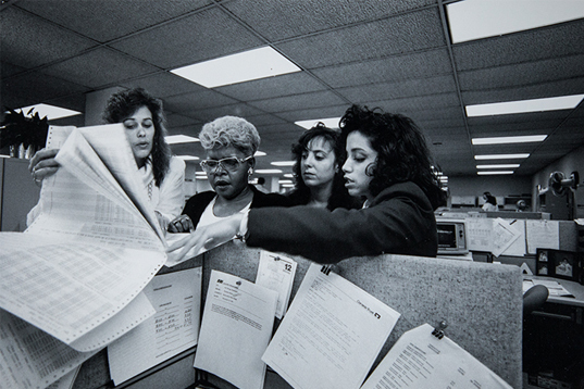 Four women reviewing paperwork at a cubicle.