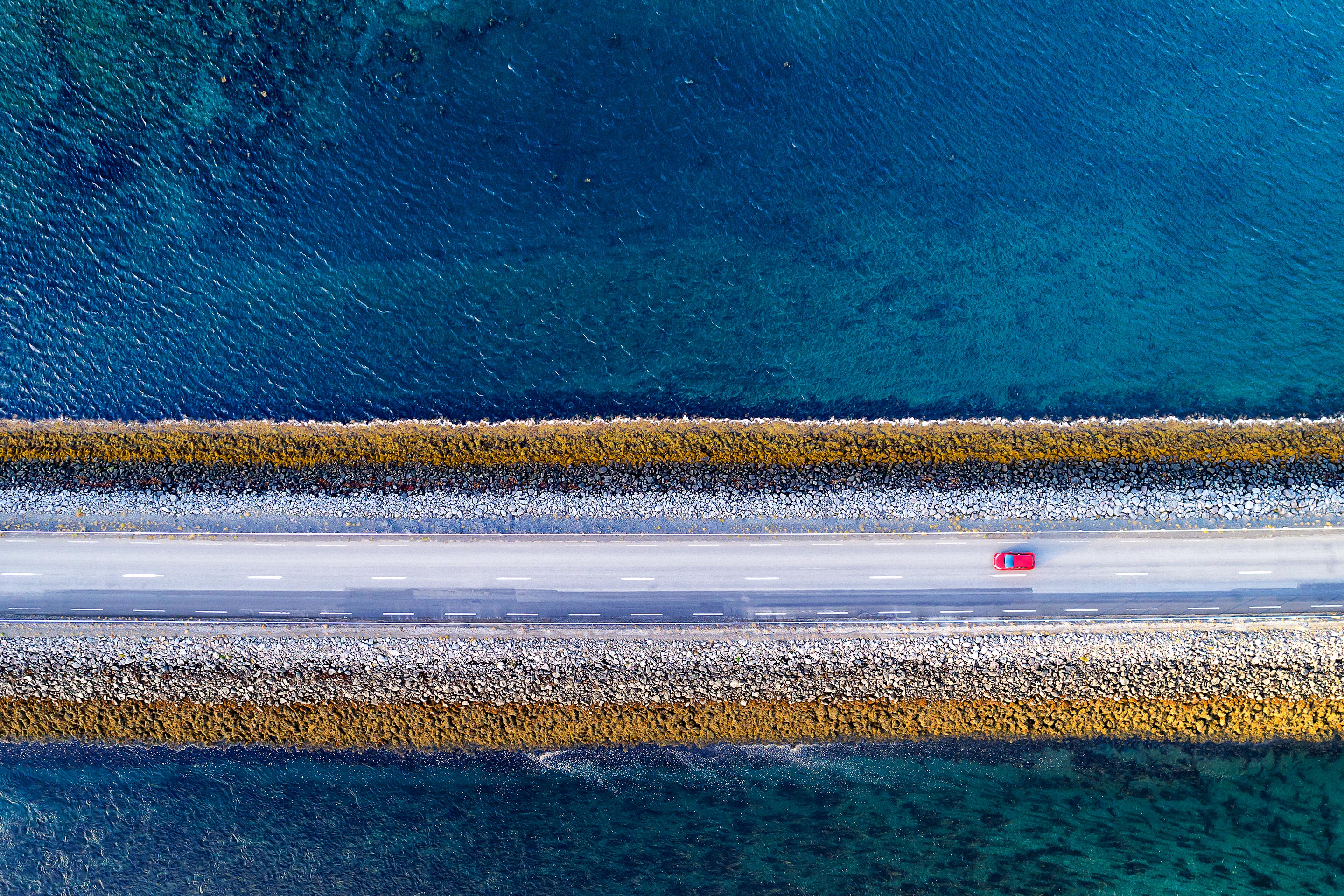 A red car drives down a single road over blue water, representing connectivity.
