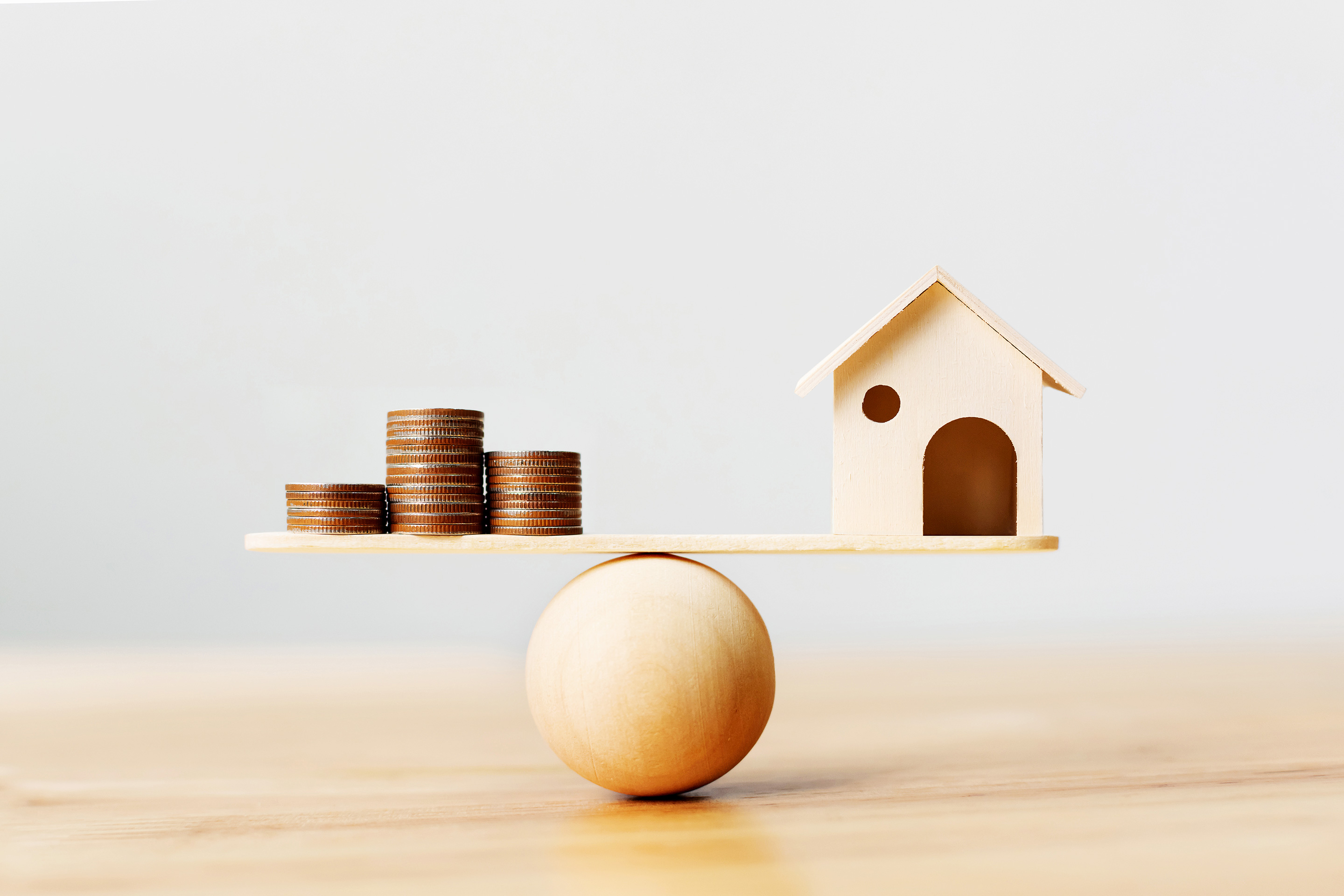 Image of a wooden ball balancing coins on one end and a small miniature wood house on the opposite end