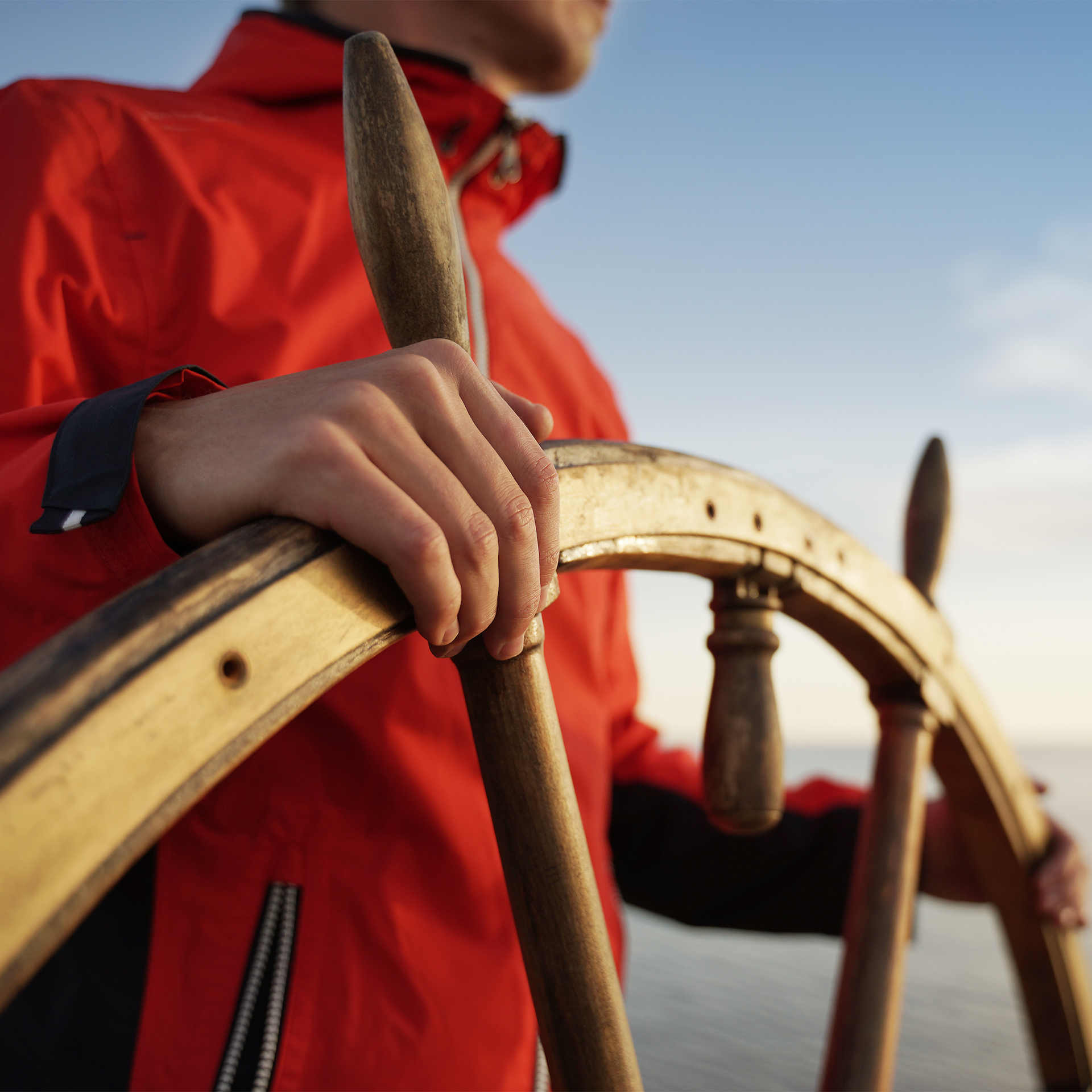 Man in a red coat steering a boat with a wooden ship wheel