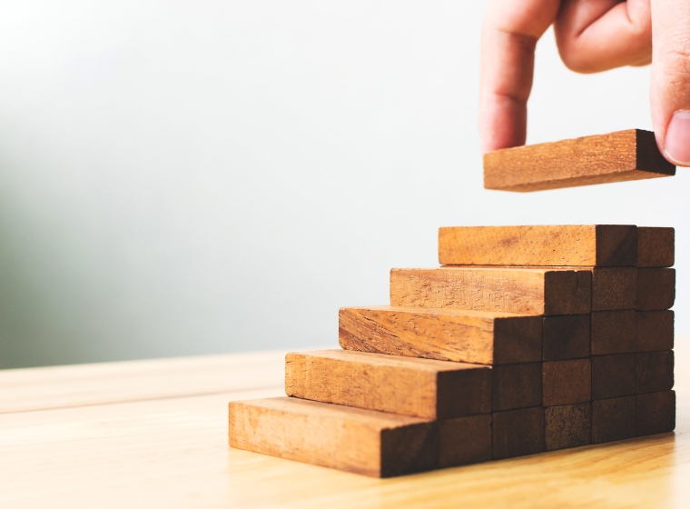 A hand adds a block to a wood puzzle in the shape of ascending stairs, as a metaphor for potential investment performance