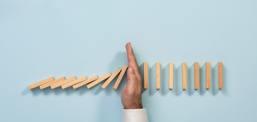 A hand divides a row of wooden blocks, stopping the domino effect representing adaptability.