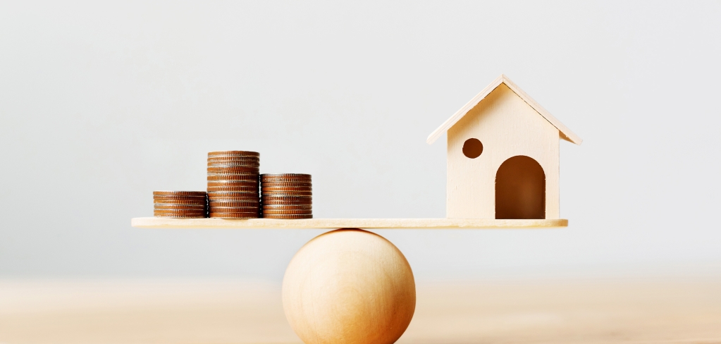 Image of a wooden ball balancing coins on one end and a small miniature wood house on the opposite end