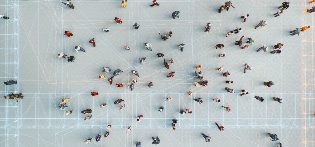 Viewed from high above, dozens of people in colorful clothing meander across a floor lined with interconnected, geometrical lines.