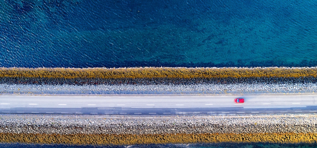 A red car drives down a single road over blue water, representing connectivity.