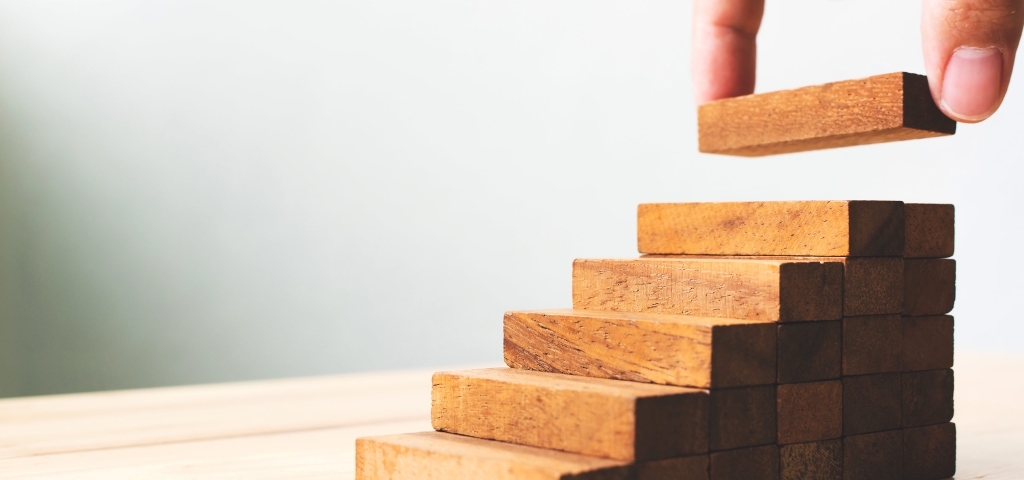 A hand adds a block to a wood puzzle in the shape of ascending stairs, as a metaphor for potential investment performance