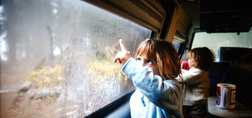 Family vacation: Two small children watch water droplets on the windows of a van, which is traveling through a forest.