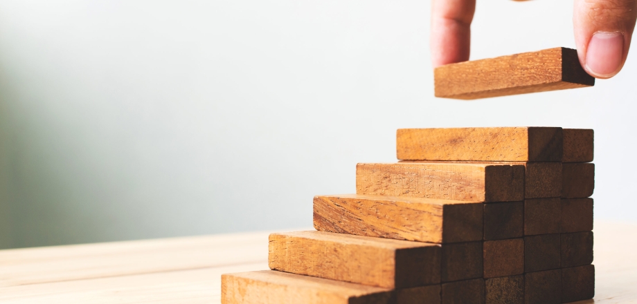 A hand adds a block to a wood puzzle in the shape of ascending stairs, as a metaphor for potential investment performance