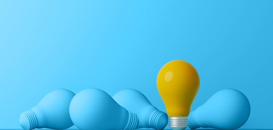 A pile of blue light bulbs lie on a blue field, looking lifeless, but one bright yellow lightbulb is standing up.