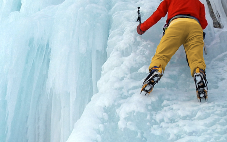 A clinber in red and yellow snow gear scales a steep cliff covered in ice. He's supported by climbing gear and boots.